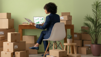 A business professional sits at a desk surrounded by stacked shipping boxes, reviewing documents while a laptop displays upward-trending sales graphs, suggesting an e-commerce or small business operation experiencing growth.